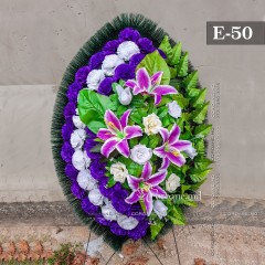 Coroana funerara originală 100cm din flori artificiale. Flori: garoafe albe, garoafe violete, crini violeţi, trandafiri albi. Frunziş: ferigă. Carcasă: cetină.